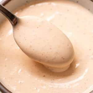 A spoon scoops up creamy, light orange Bang Bang Sauce with specks, from a ceramic bowl.