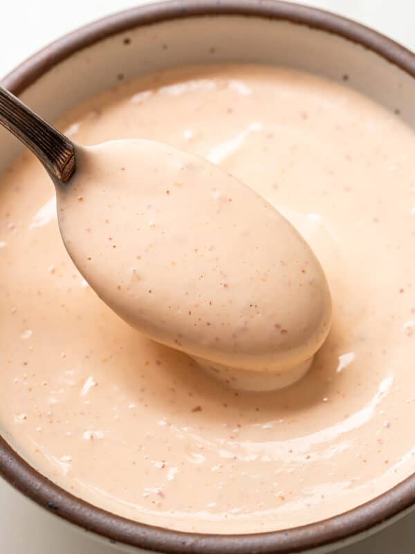 A spoon scoops up creamy, light orange Bang Bang Sauce with specks, from a ceramic bowl.