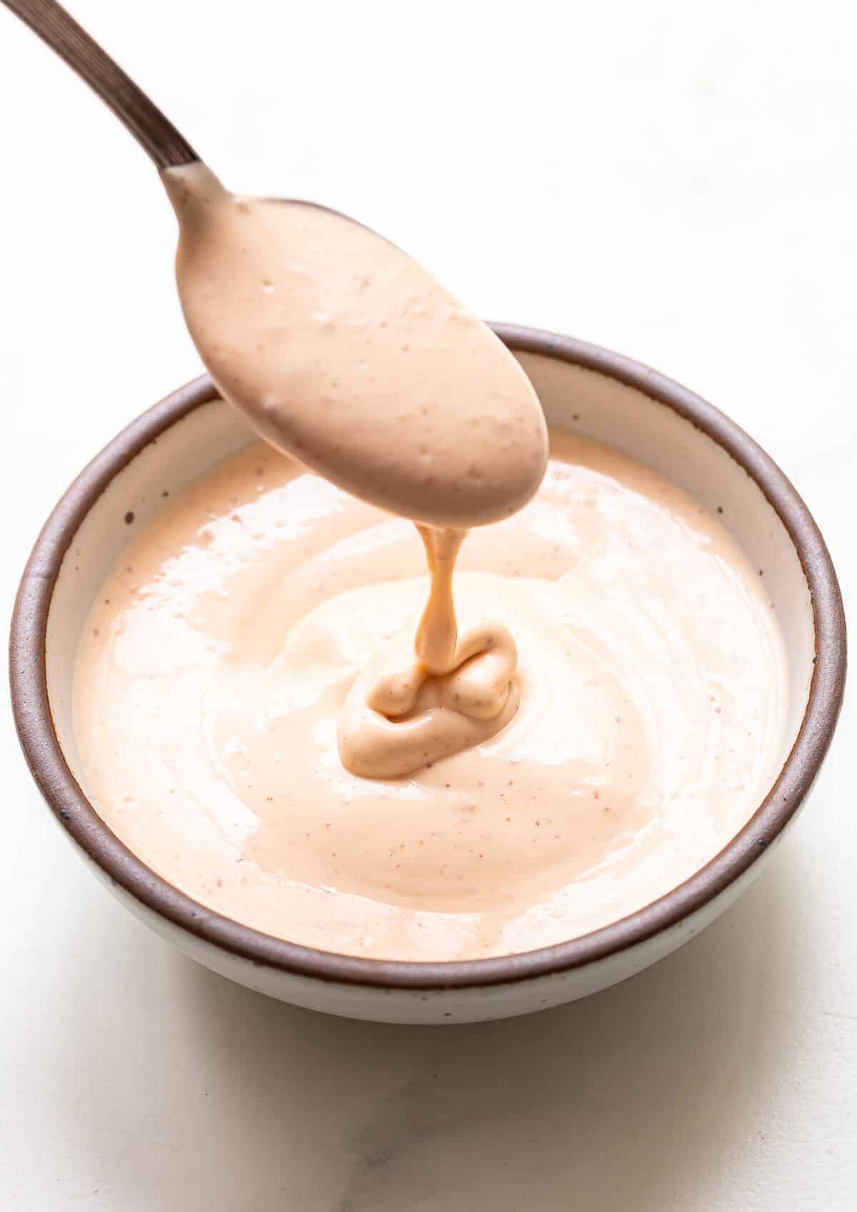 A spoon drizzling creamy, light orange Bang Bang Sauce into a small bowl filled with the same sauce, on a white surface.