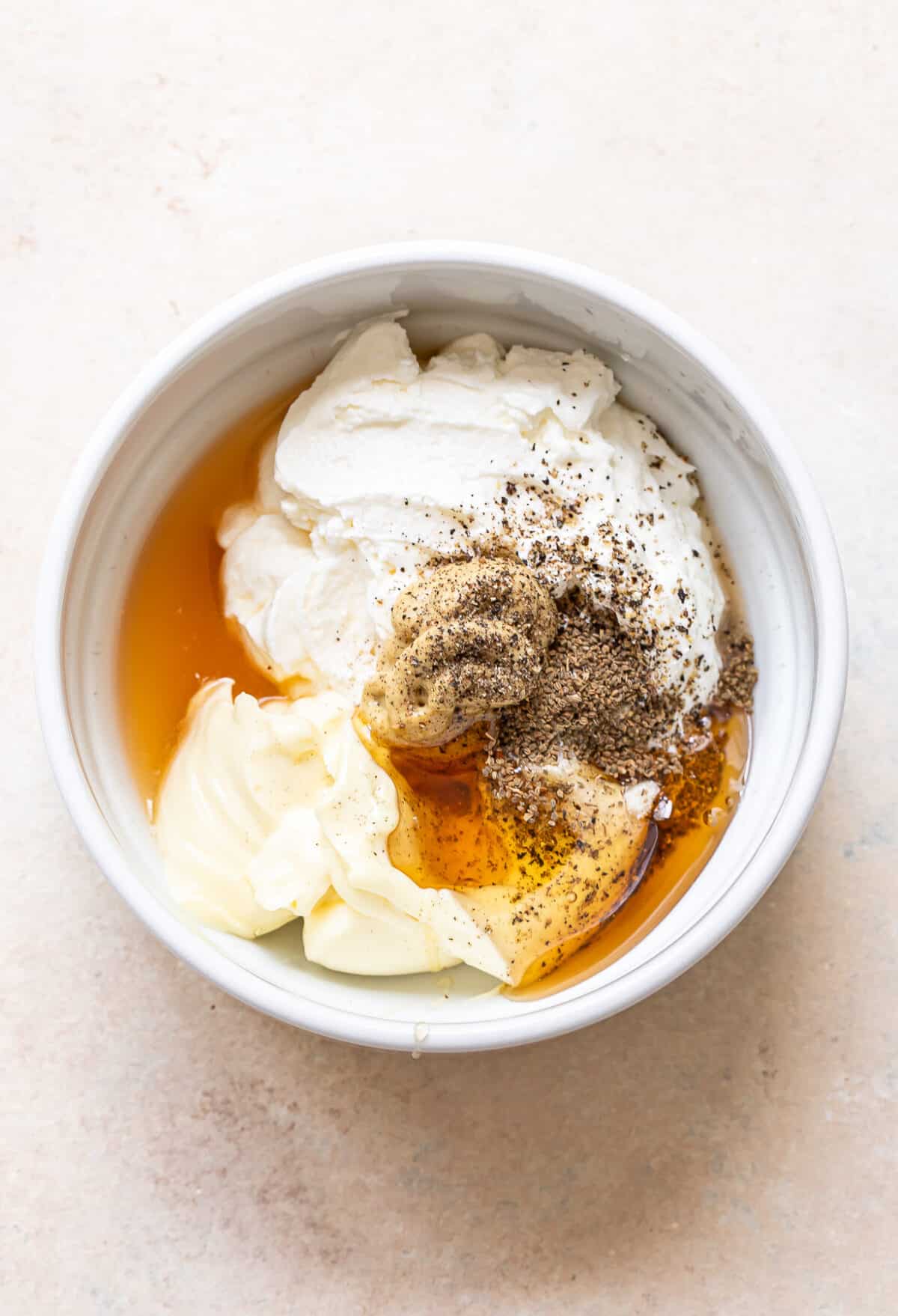 A white bowl contains the creamy base for coleslaw—yogurt, mayonnaise, honey, Dijon mustard, and cracked black pepper—ready to be mixed.
