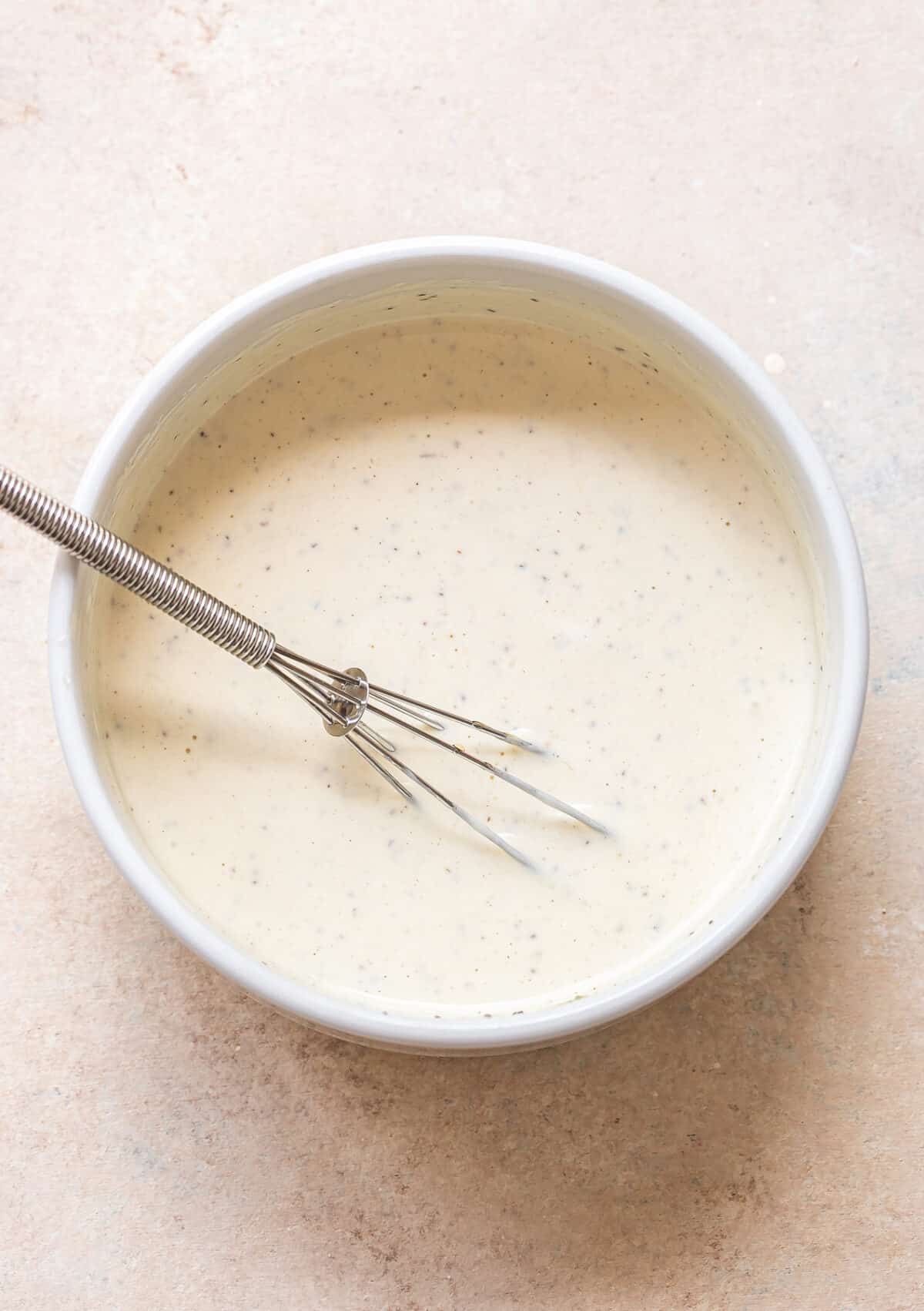 A metal whisk rests in a white bowl filled with creamy, speckled coleslaw dressing or sauce, placed on a light beige surface.