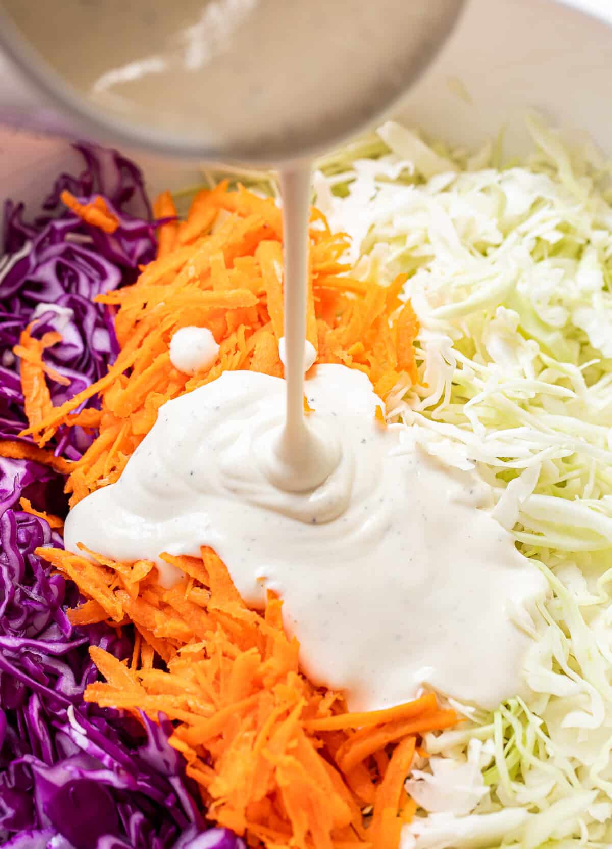 Creamy dressing being poured over shredded purple cabbage, carrots, and green cabbage in a bowl, ready to be mixed into a classic coleslaw.
