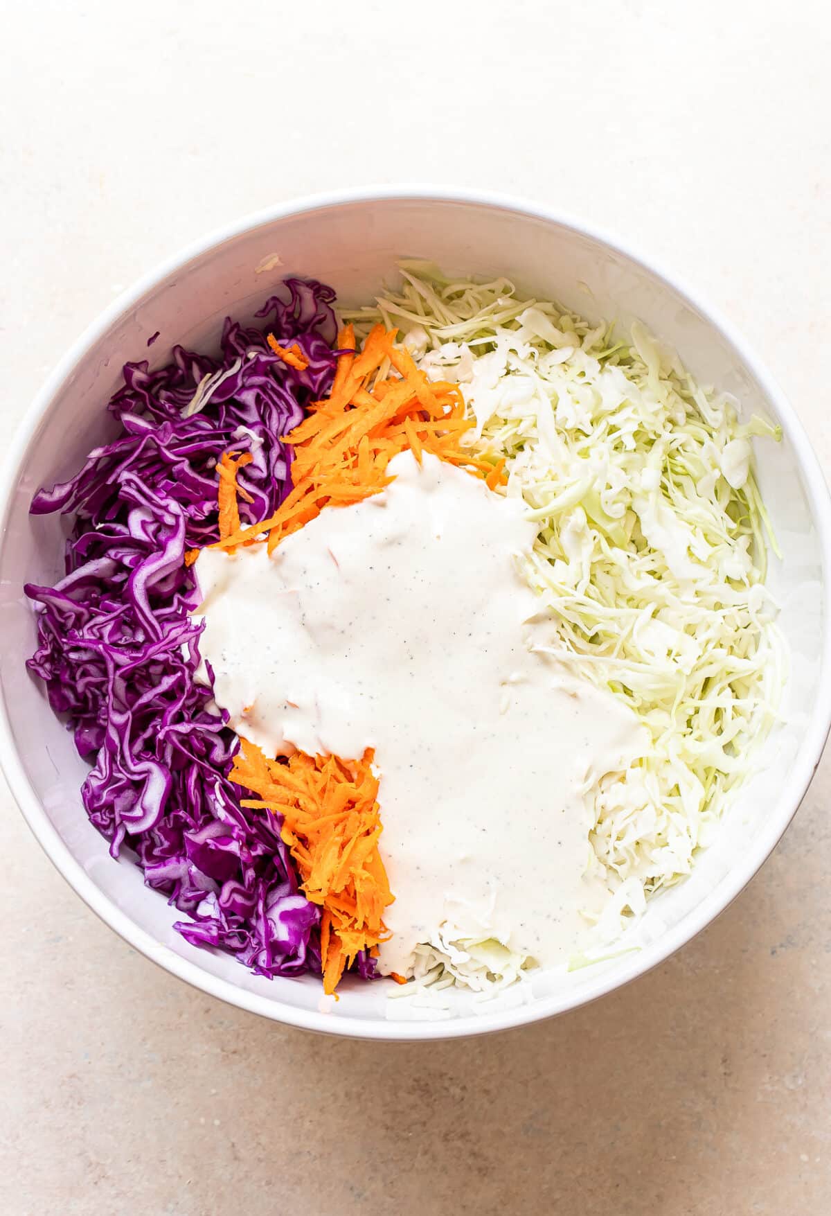 A bowl containing shredded purple cabbage, shredded green cabbage, grated carrots, and a generous portion of creamy dressing on top, ready to be mixed for classic coleslaw.
