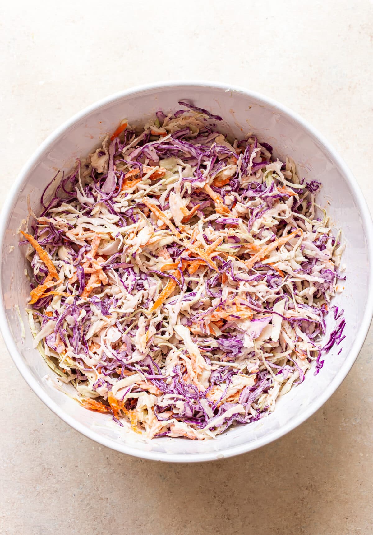 A white bowl filled with flavorful coleslaw made of shredded green and purple cabbage, with orange carrot strips, all mixed with a creamy dressing, sits on a light countertop.
