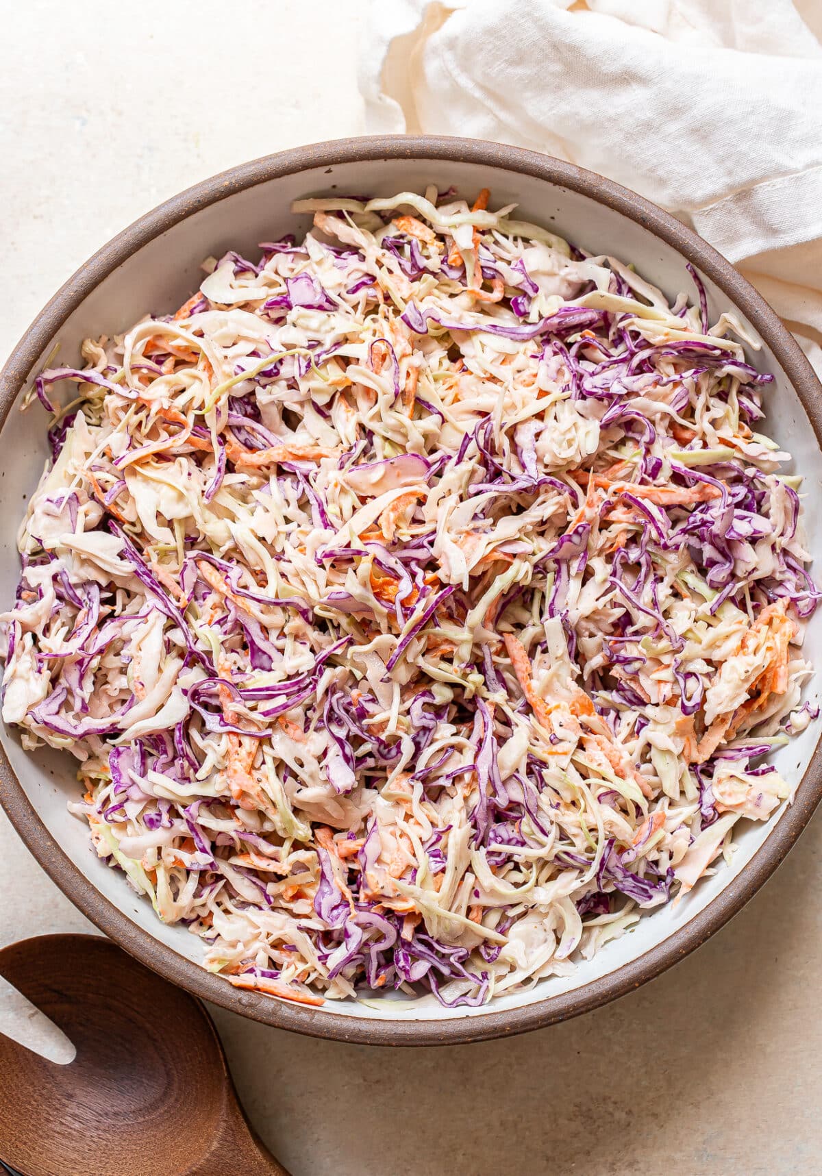 A bowl of creamy coleslaw with shredded green and purple cabbage and carrots is the star of this fresh scene. A wooden spoon and a white cloth are placed beside the coleslaw bowl, ready for serving.