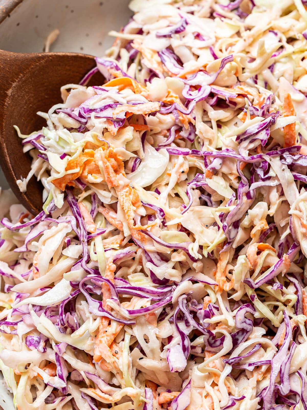 Close-up of creamy coleslaw with shredded green and purple cabbage and carrots being mixed with a wooden spoon, highlighting the fresh ingredients that make this classic coleslaw so vibrant and delicious.