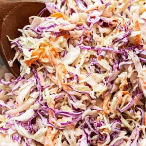 Close-up of creamy coleslaw with shredded green and purple cabbage and carrots being mixed with a wooden spoon, highlighting the fresh ingredients that make this classic coleslaw so vibrant and delicious.