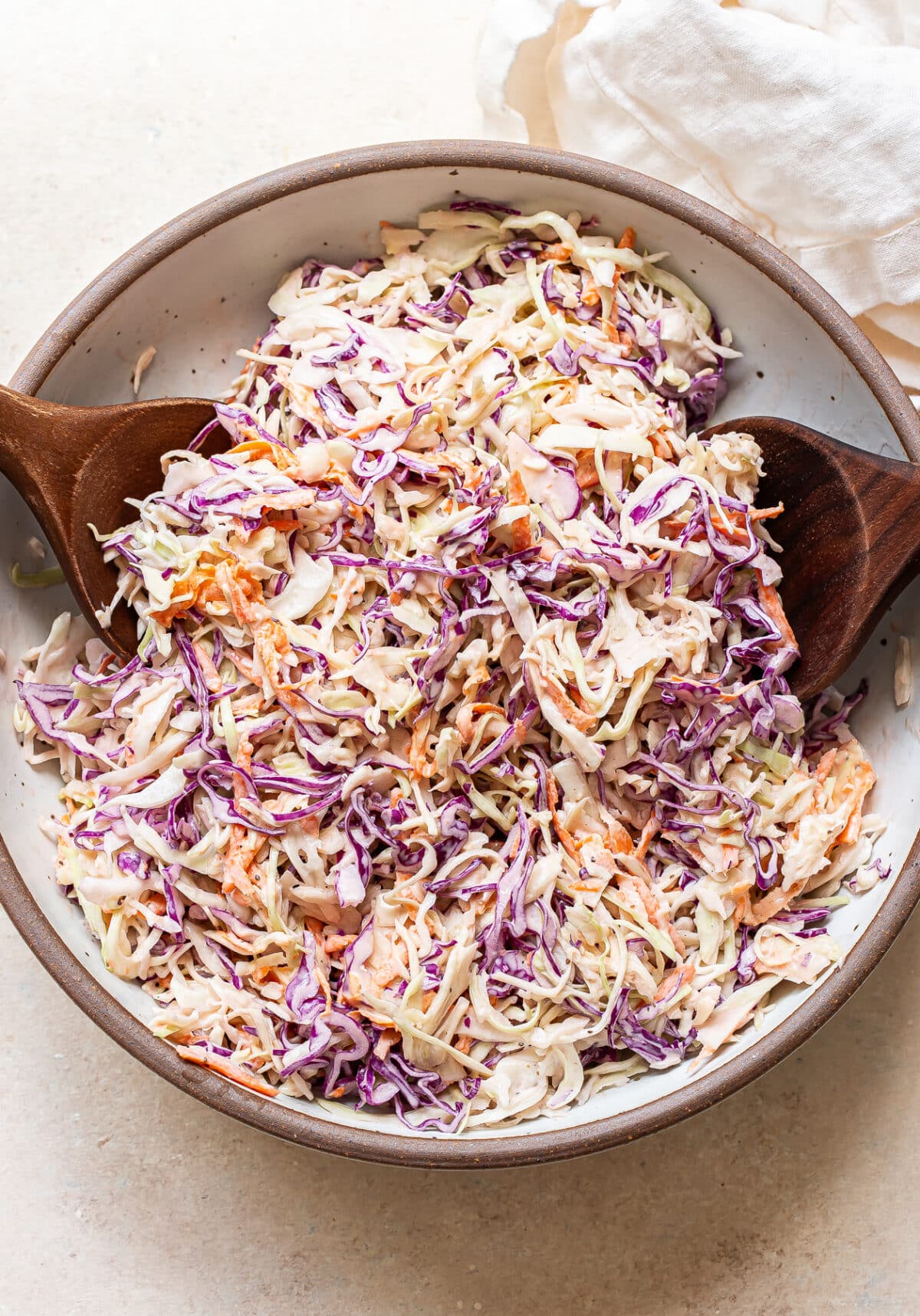 A vibrant bowl of coleslaw, featuring shredded green and purple cabbage and carrots tossed in a creamy dressing. Two wooden serving spoons rest in the bowl, ready for serving. A white cloth napkin lies nearby, completing this fresh coleslaw presentation.