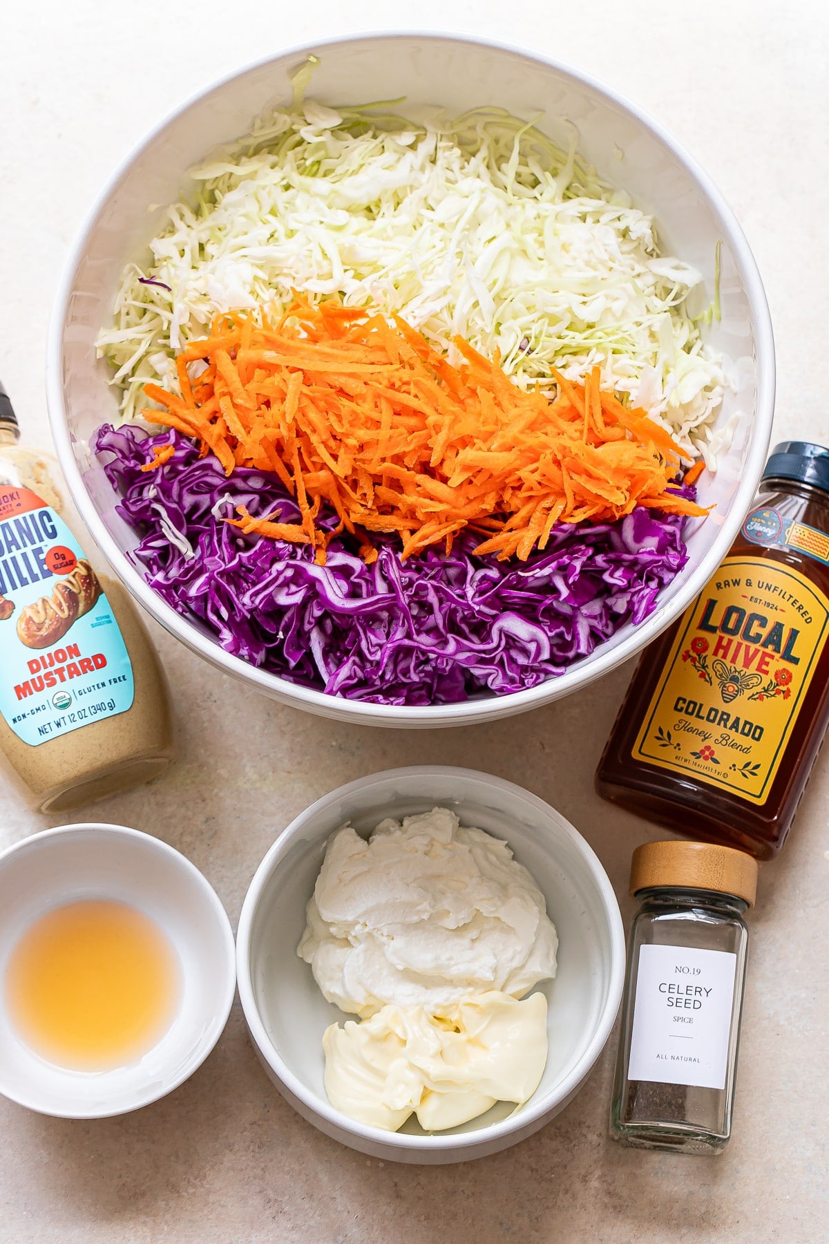 A bowl of crisp coleslaw with shredded green cabbage, purple cabbage, and carrots stands at the center. Surrounding it are Dijon mustard, honey, celery seed, mayonnaise, and a small bowl of apple cider vinegar—perfect ingredients for crafting a classic coleslaw dressing.