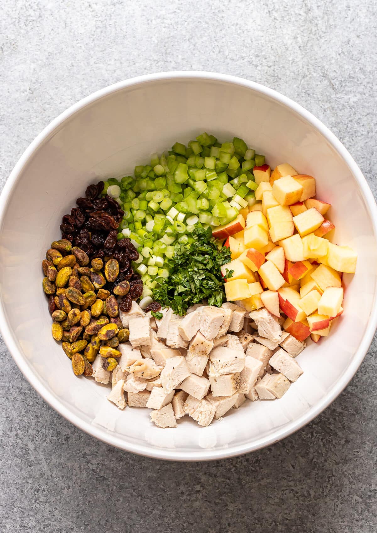 A white bowl with Curry Chicken Salad featuring diced chicken, apples, celery, raisins, pistachios, and chopped herbs, all arranged in separate sections on a gray surface.