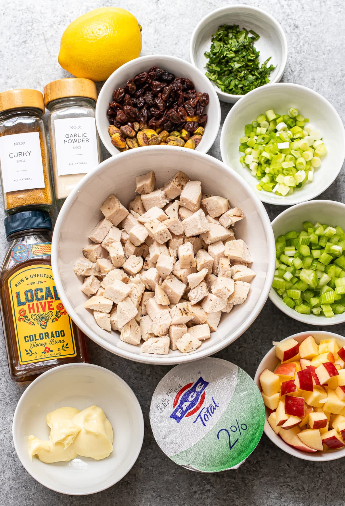 Bowls of diced cooked chicken, chopped apples, celery, green onions, raisins, parsley, curry powder, garlic powder, honey, mayonnaise, Greek yogurt, and a lemon on a gray surface—everything you need to create a vibrant and flavorful Curry Chicken Salad.