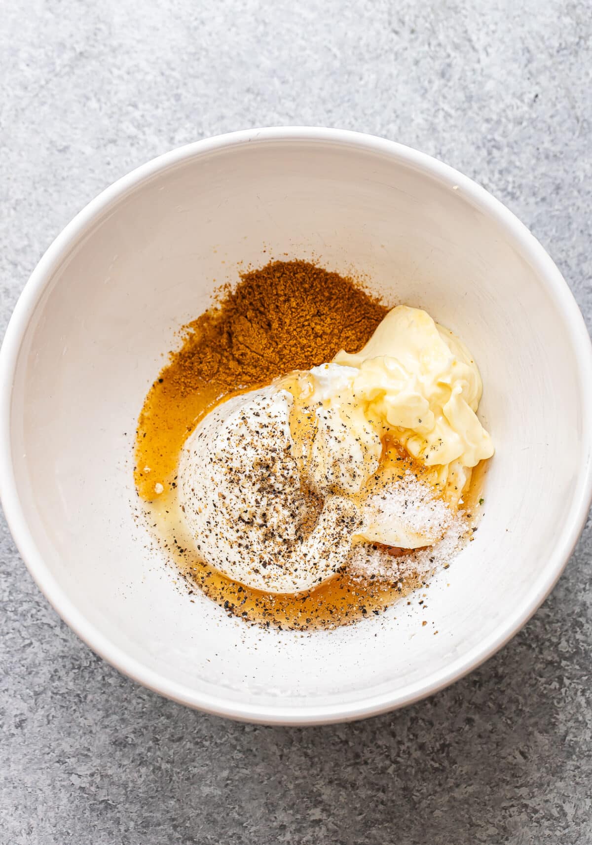 A white bowl containing mayonnaise, sour cream, spices, ground pepper, and a brown seasoning mix—perfect for making a flavorful Curry Chicken Salad—sits on a gray surface.