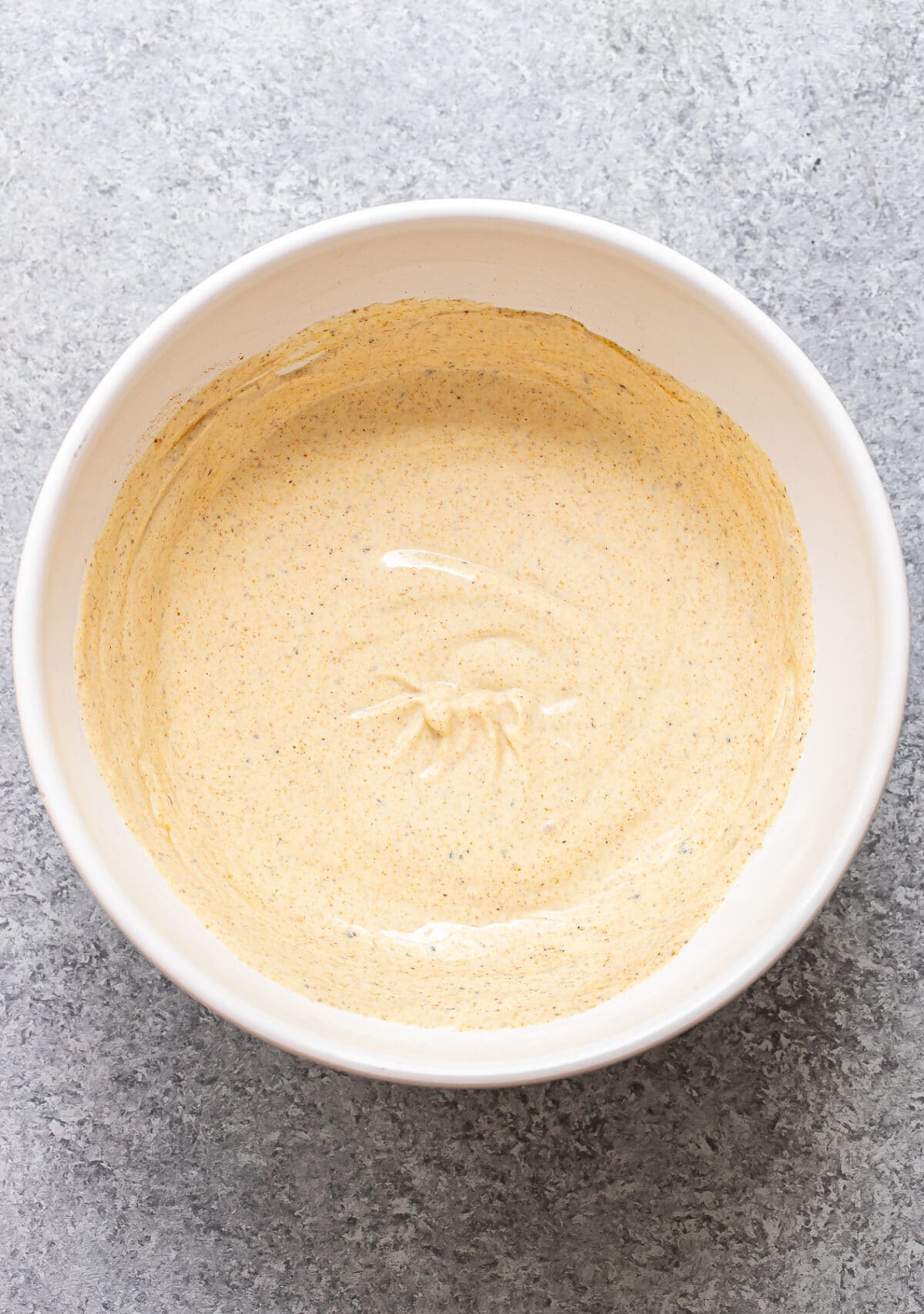 A white bowl filled with a smooth, light brown batter sits on a gray textured surface. Reminiscent of the warm hues found in a Curry Chicken Salad, the batter appears evenly mixed, with a few small bubbles visible on the surface.