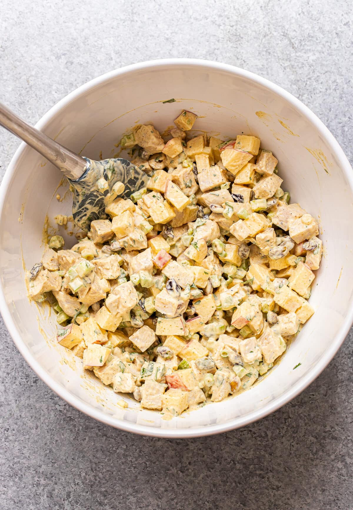 A white bowl filled with a creamy Curry Chicken Salad-inspired mixture of cubed tofu, chopped celery, apples, and herbs, being mixed with a spatula on a gray countertop.