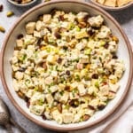 A bowl of Curry Chicken Salad with chopped apples, celery, pistachios, dried cranberries, and herbs, served with a side of crackers. Two spoons and a napkin are nearby.