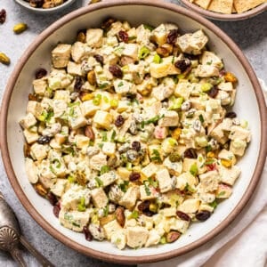 A bowl of Curry Chicken Salad with chopped apples, celery, pistachios, dried cranberries, and herbs, served with a side of crackers. Two spoons and a napkin are nearby.