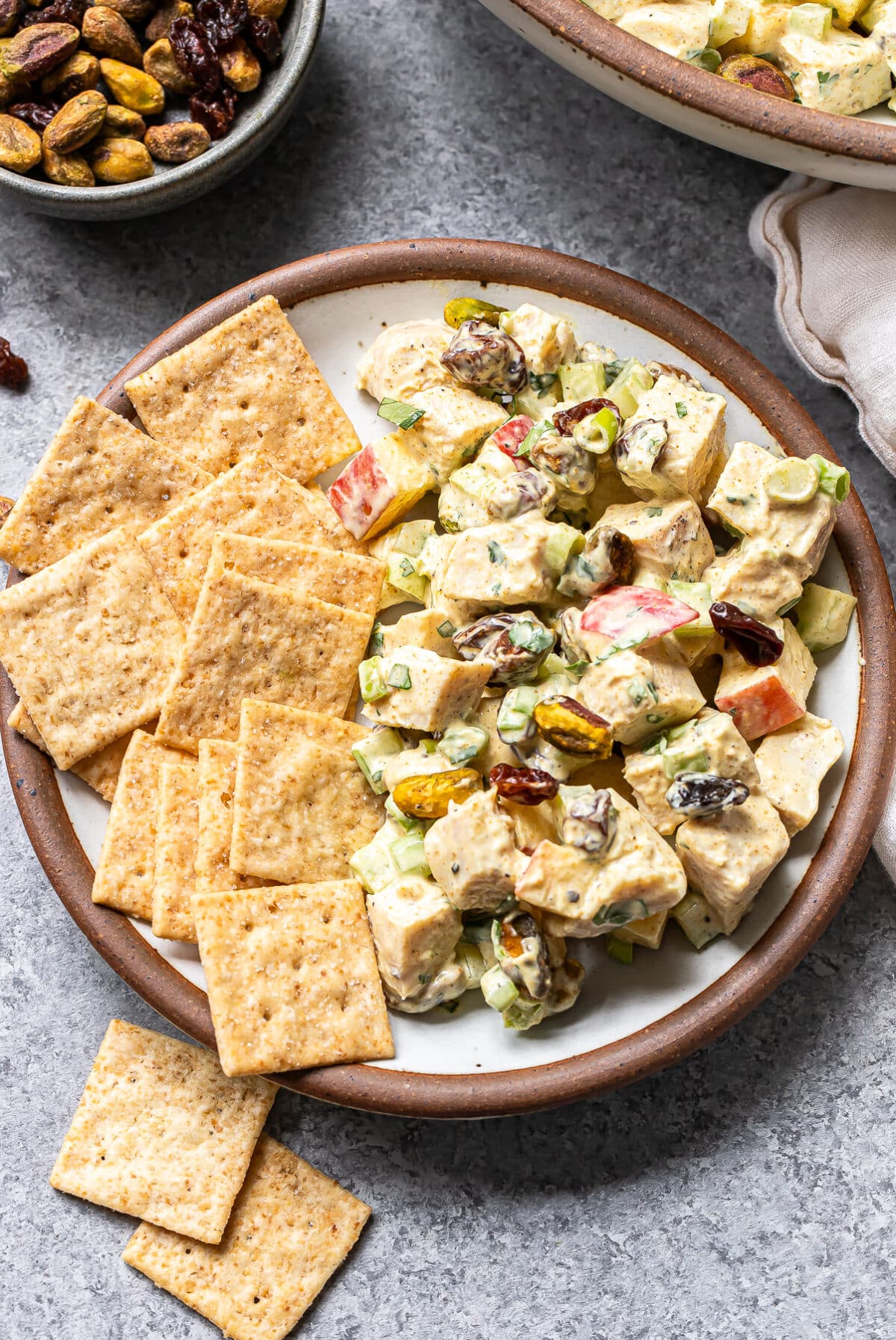 A plate with crackers and a flavorful Curry Chicken Salad containing apples, celery, raisins, and pistachios in a creamy dressing. A bowl of nuts and part of another dish are visible nearby.