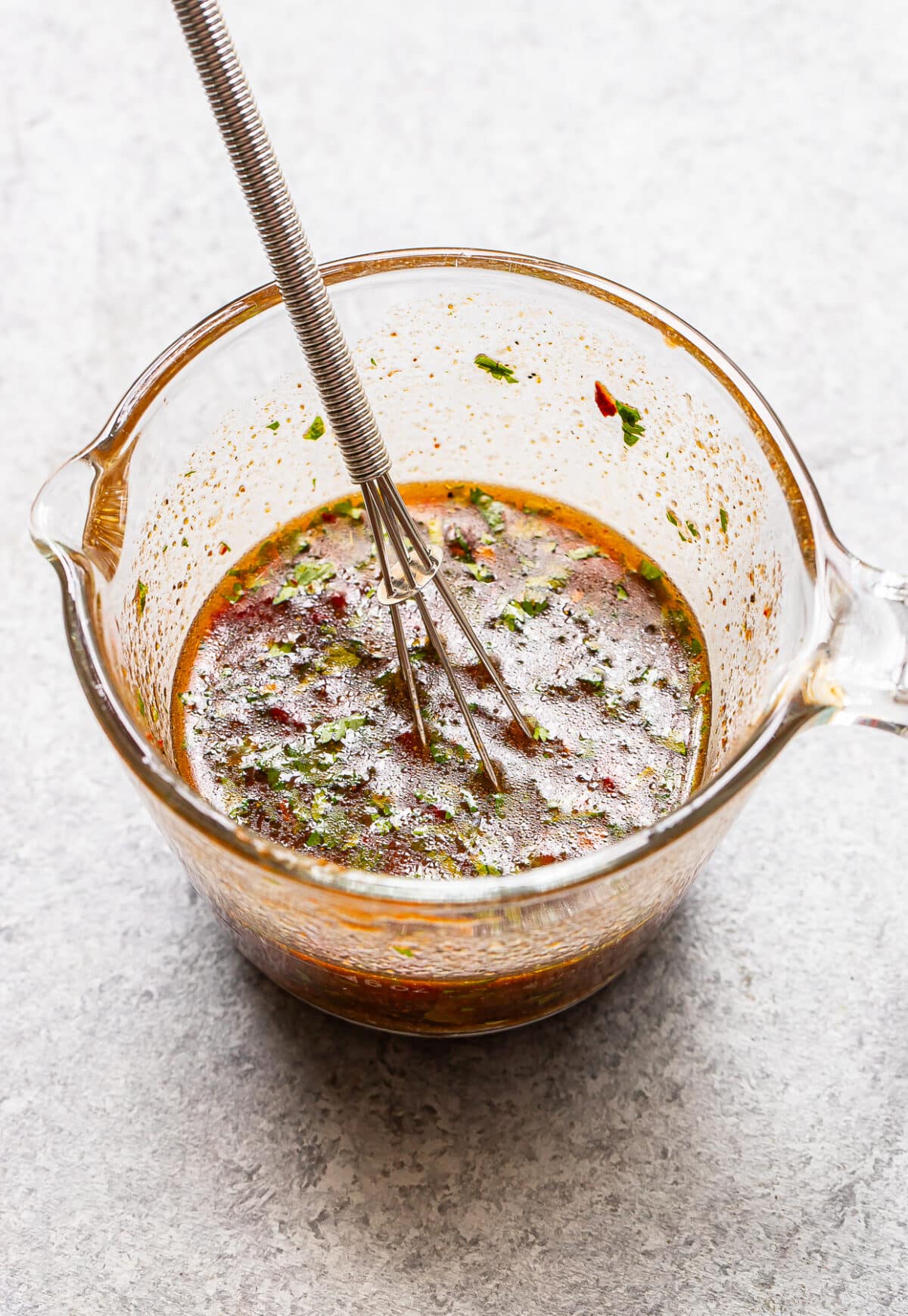 A metal whisk rests in a glass measuring cup filled with a reddish-brown, herbed liquid mixture&mdash;perfect for marinating Honey Chipotle Chicken&mdash;set on a light gray countertop.