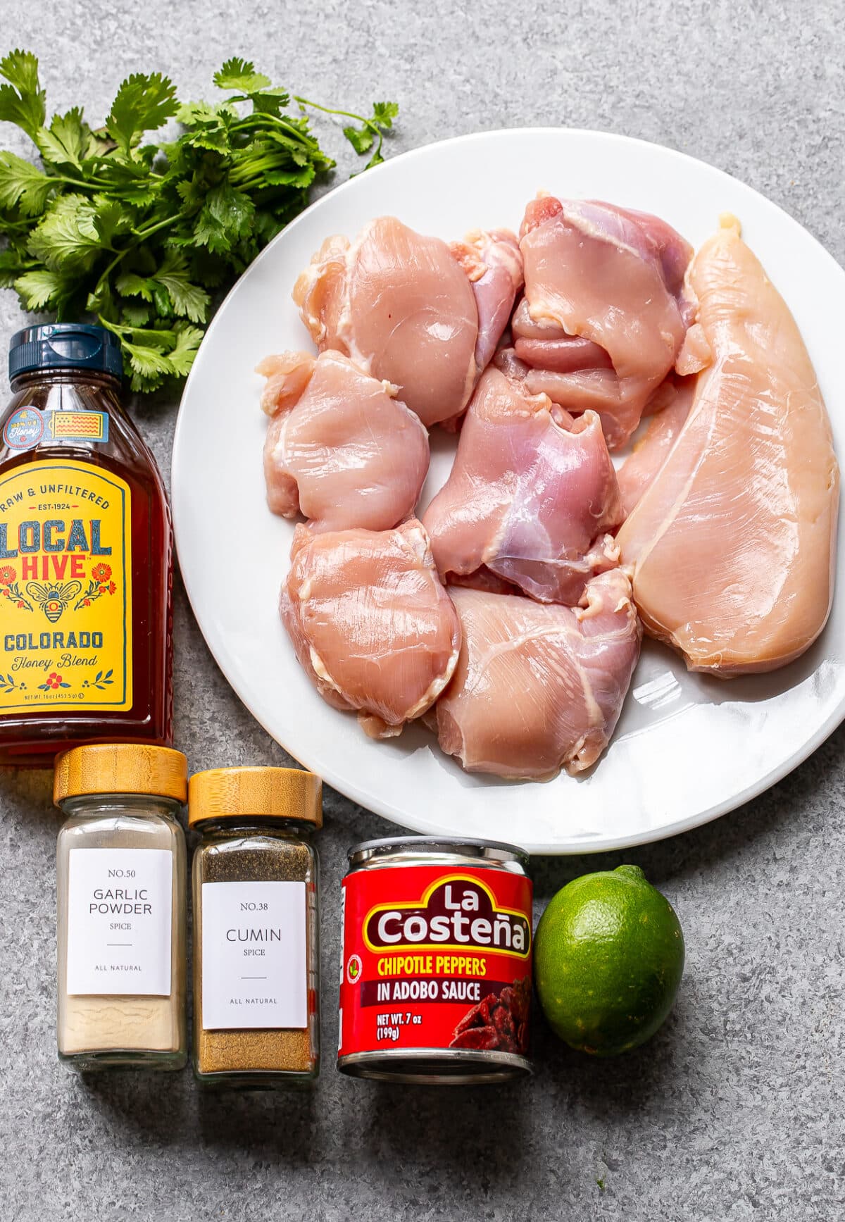 Raw chicken pieces on a white plate, with cilantro, honey, garlic powder, cumin, chipotle peppers in adobo sauce, and a lime arranged on a gray surface&mdash;perfect for making delicious Honey Chipotle Chicken.