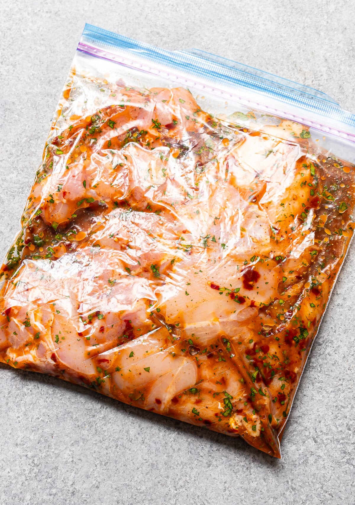 Raw chicken marinating in a clear plastic zip-top bag with herbs and spices for a flavorful Honey Chipotle Chicken, placed on a light gray surface.