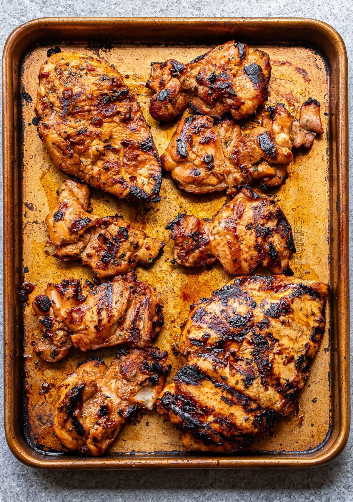 Grilled, marinated Honey Chipotle Chicken pieces with char marks arranged on a well-used baking sheet, viewed from above on a light surface.