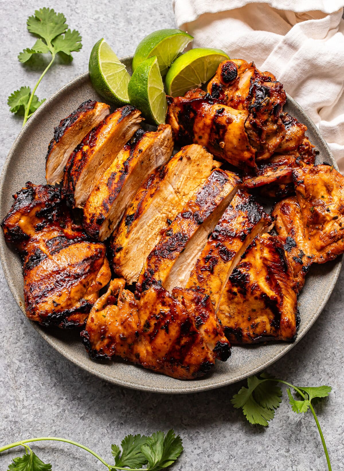 A plate of grilled, sliced Honey Chipotle Chicken thighs with char marks, garnished with lime wedges and fresh cilantro leaves, sits on a gray surface next to a beige napkin.