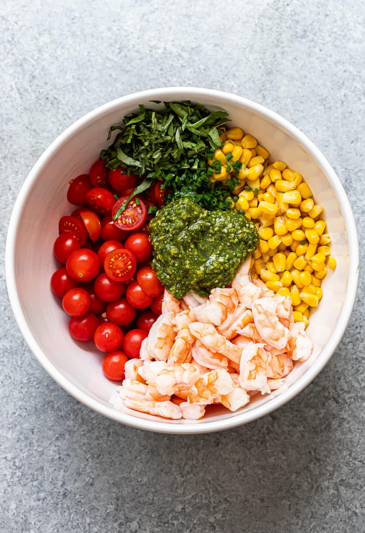 A white bowl filled with Tomato Corn Shrimp Salad—succulent shrimp, cherry tomatoes, corn, chopped herbs, and a dollop of green pesto—arranged in separate sections on a gray surface.