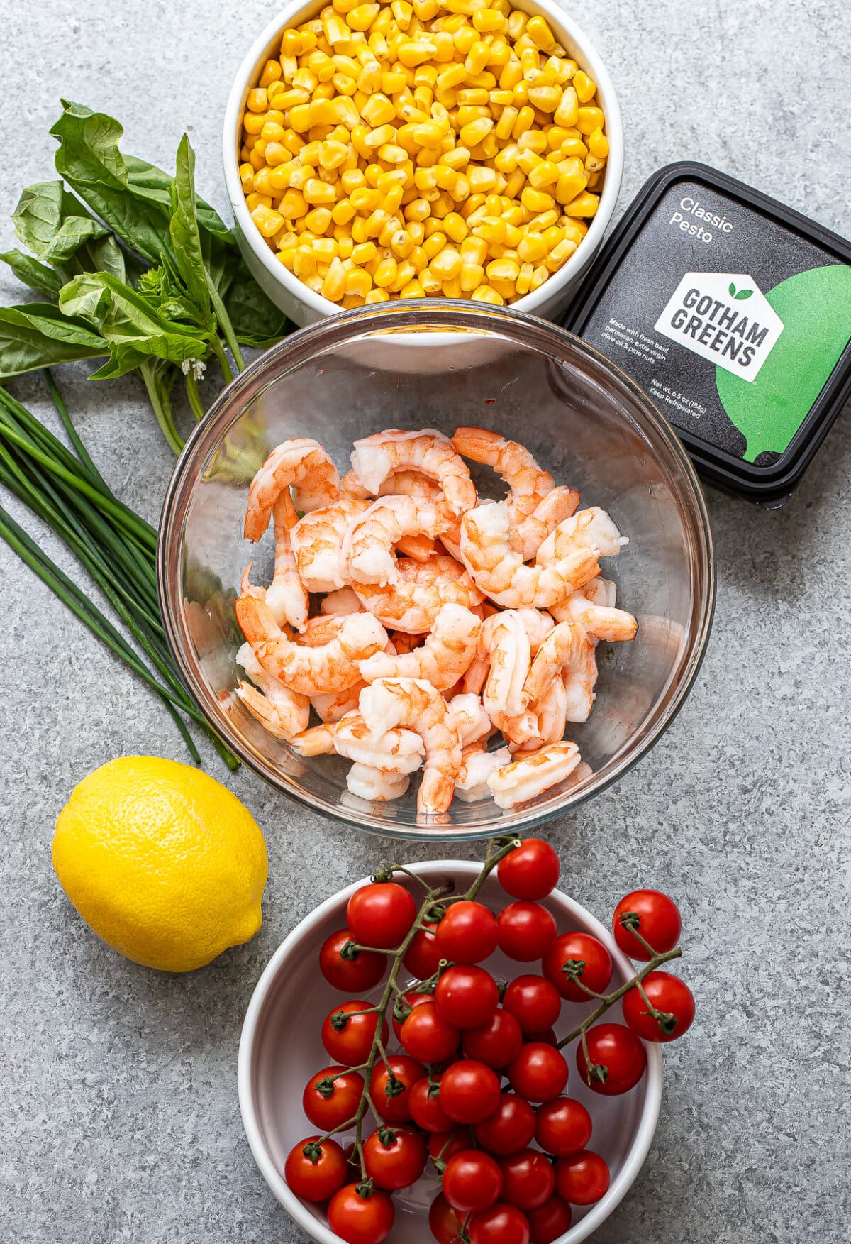 A bowl of cooked shrimp, a bowl of corn, grape tomatoes on the vine, fresh basil, chives, a lemon, and a container of classic pesto are artfully arranged on a gray surface—perfect ingredients for preparing a vibrant Tomato Corn Shrimp Salad.