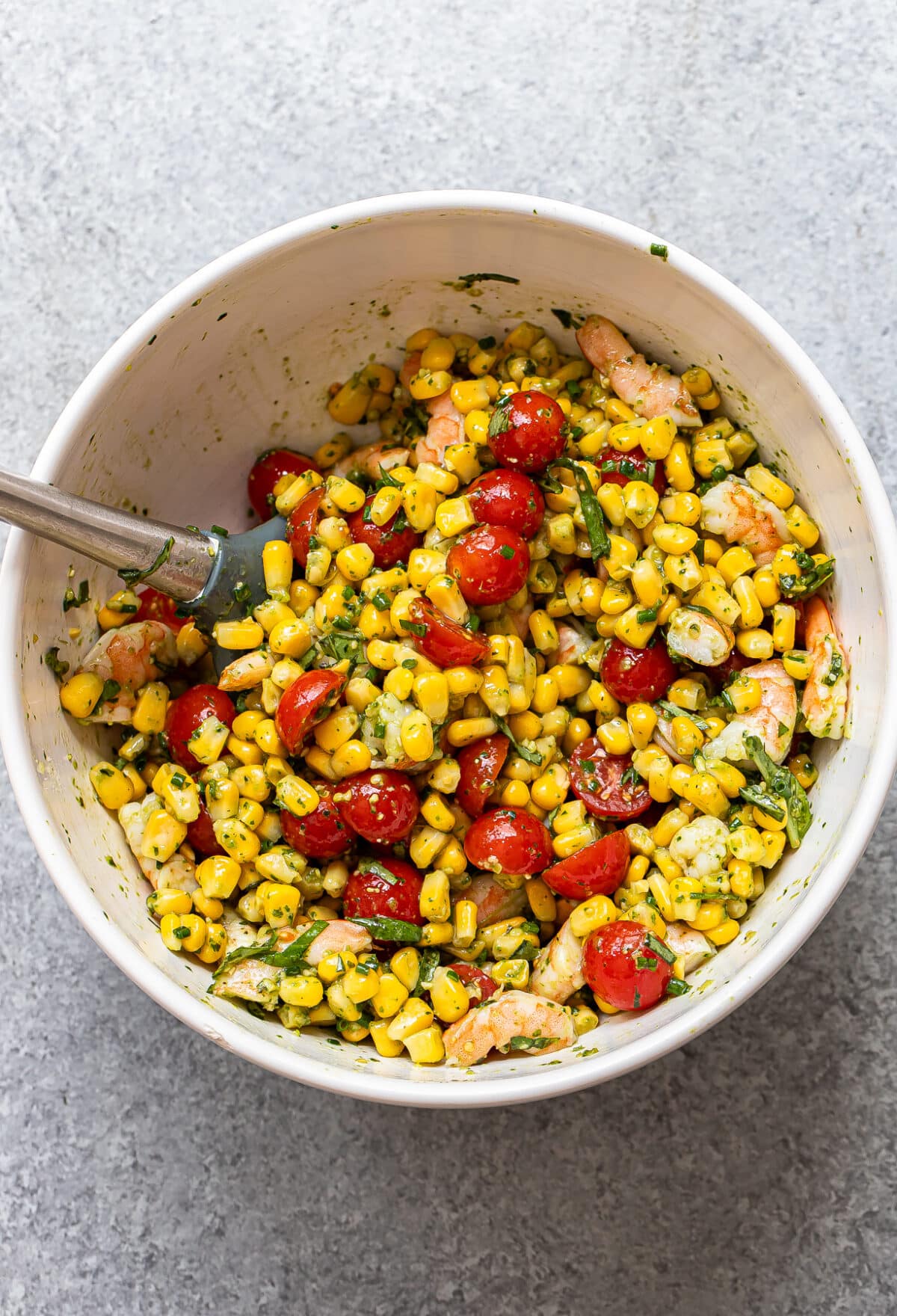 A bowl of Tomato Corn Shrimp Salad with cherry tomatoes, corn, shrimp, and chopped herbs, all mixed with a flavorful dressing. A metal spoon rests inside the bowl on a light gray surface.