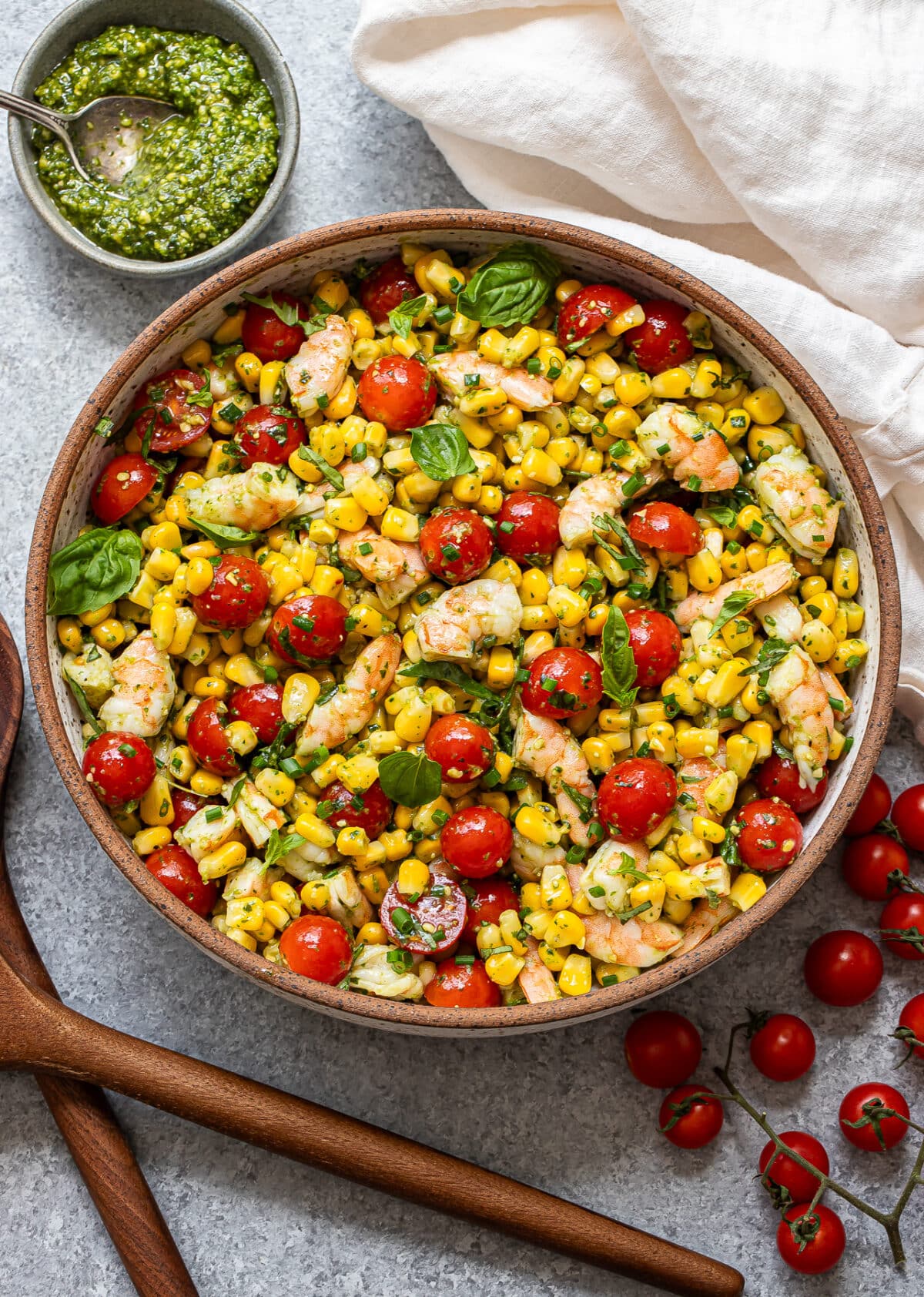 A bowl of Tomato Corn Shrimp Salad with cherry tomatoes, basil, and chives sits on a gray surface, accompanied by a small bowl of green sauce, a white napkin, and wooden salad servers nearby.