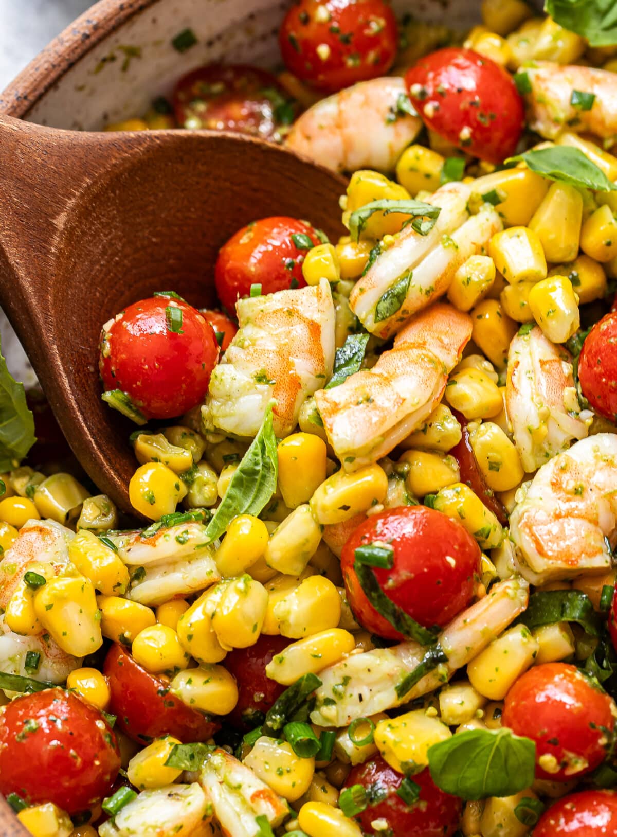 A close-up of a wooden spoon scooping a vibrant Tomato Corn Shrimp Salad brimming with shrimp, corn, cherry tomatoes, fresh basil, and herbs in a bowl.
