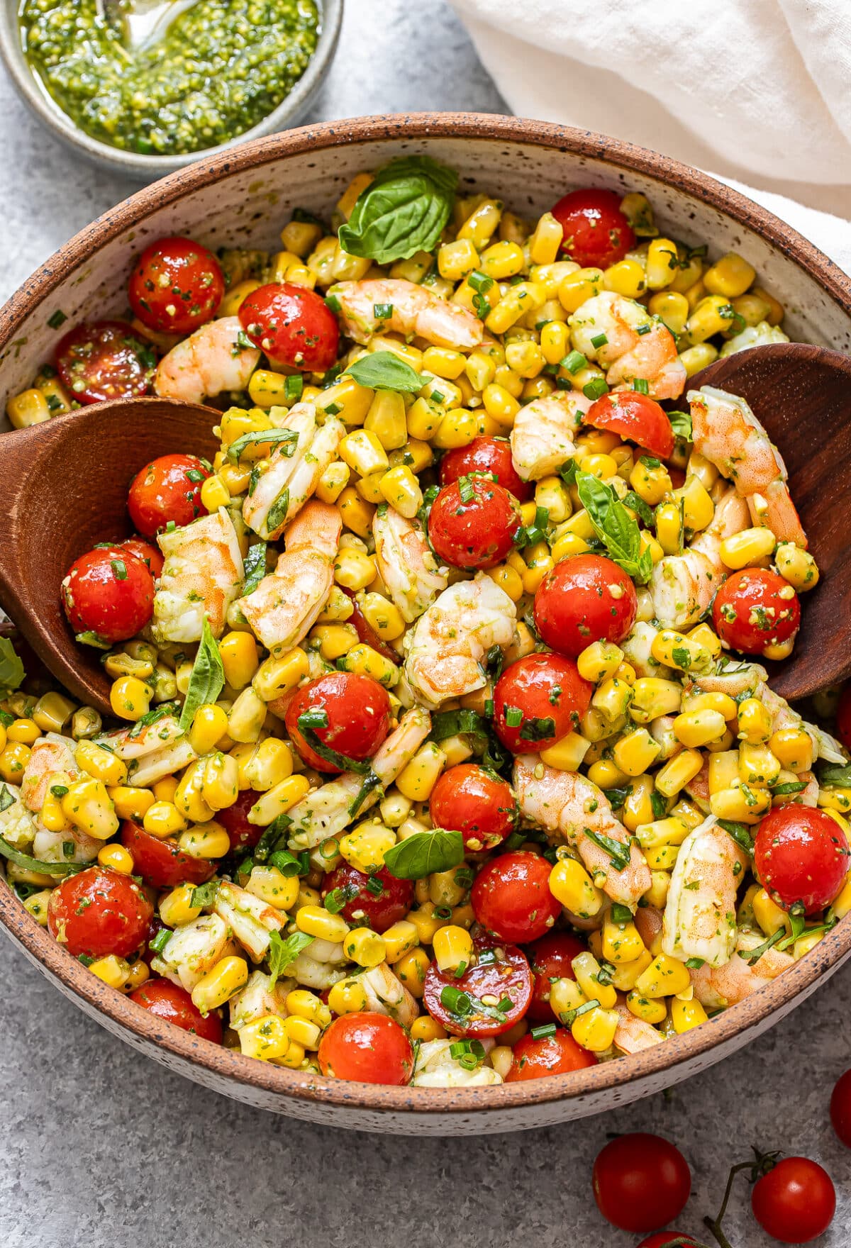 A vibrant Tomato Corn Shrimp Salad is presented in a bowl, featuring plump shrimp, juicy cherry tomatoes, sweet corn, and fresh herbs, accompanied by two wooden spoons. A small bowl of pesto and a white napkin are placed nearby.