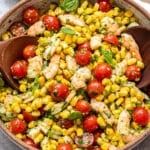 A vibrant Tomato Corn Shrimp Salad is presented in a bowl, featuring plump shrimp, juicy cherry tomatoes, sweet corn, and fresh herbs, accompanied by two wooden spoons. A small bowl of pesto and a white napkin are placed nearby.