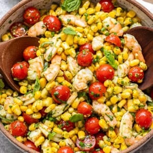 A vibrant Tomato Corn Shrimp Salad is presented in a bowl, featuring plump shrimp, juicy cherry tomatoes, sweet corn, and fresh herbs, accompanied by two wooden spoons. A small bowl of pesto and a white napkin are placed nearby.