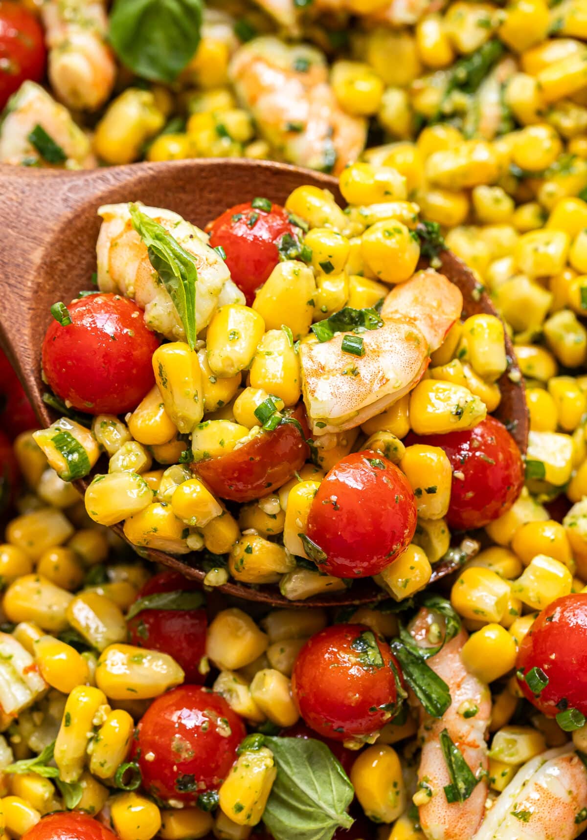 A wooden spoon scoops Tomato Corn Shrimp Salad with cherry tomatoes, basil leaves, and herbs, all mixed with a light dressing.