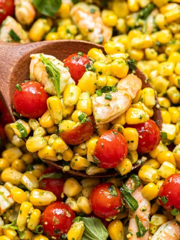 A wooden spoon scoops Tomato Corn Shrimp Salad with cherry tomatoes, basil leaves, and herbs, all mixed with a light dressing.