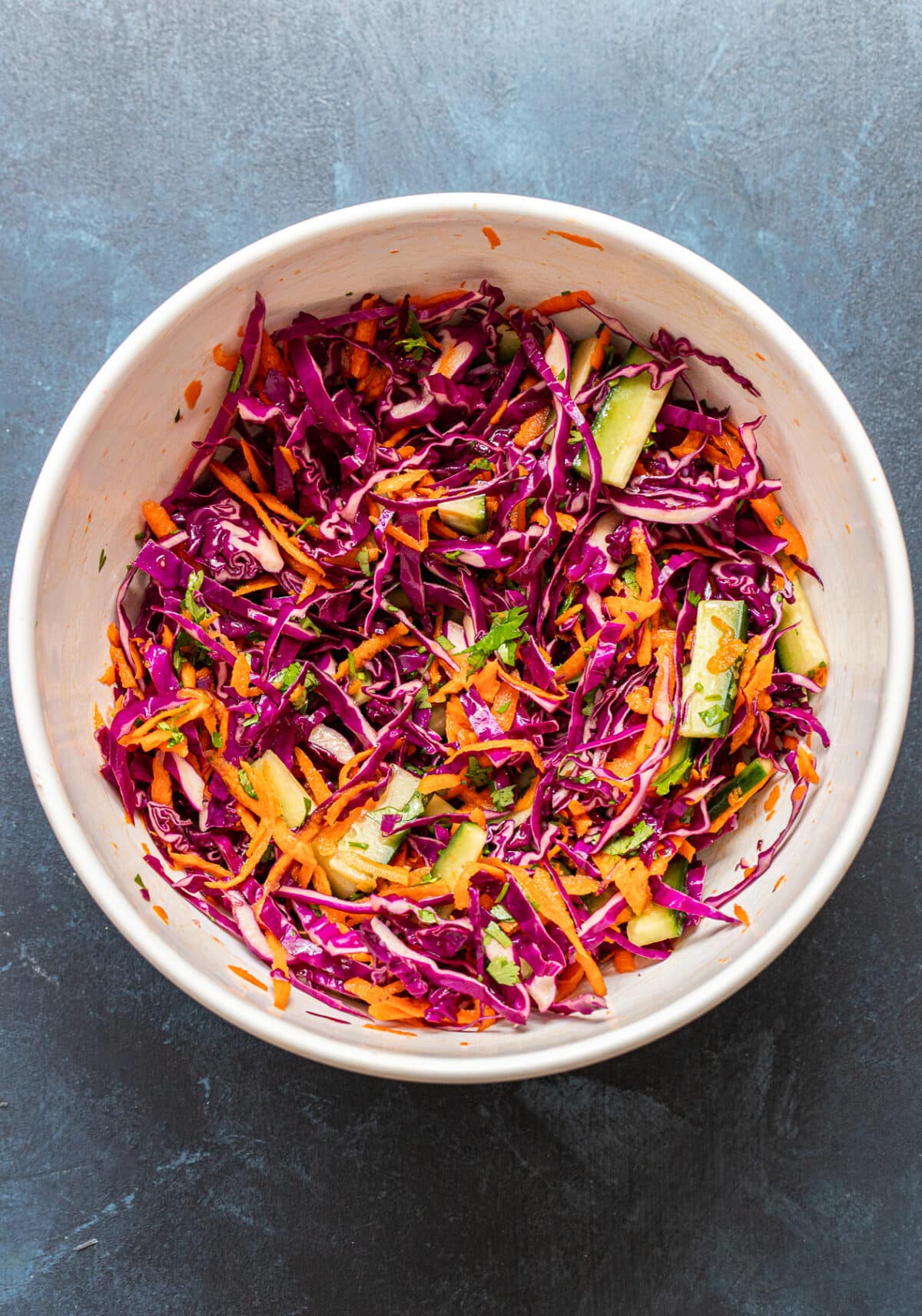A white bowl filled with colorful coleslaw—made of shredded red cabbage, carrots, cucumbers, and herbs—sits on a dark gray surface, making the perfect fresh accompaniment to Bang Bang Chicken Bowls.