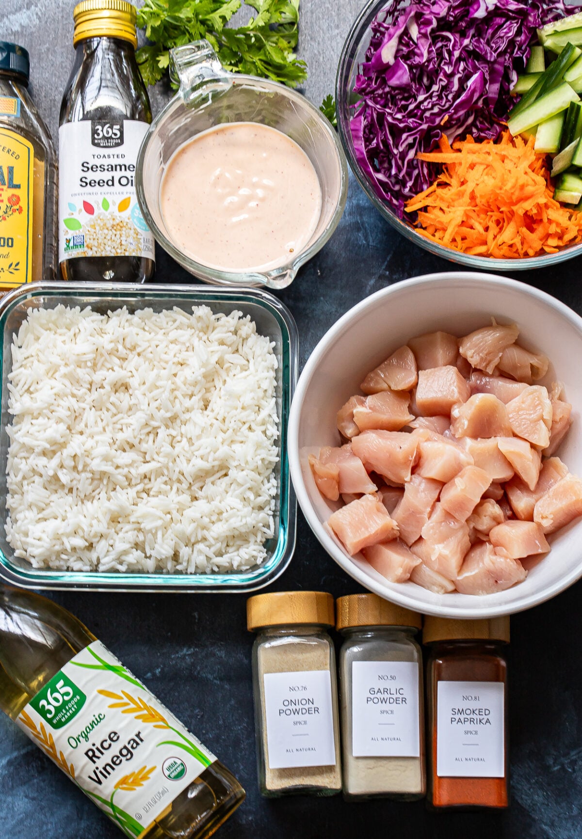 Chopped raw chicken, cooked rice, shredded cabbage, carrots, cilantro, a creamy sauce, rice vinegar, sesame oil, and jars of onion powder, garlic powder, and smoked paprika are artfully arranged on a dark surface—everything you need to create delicious Bang Bang Chicken Bowls.