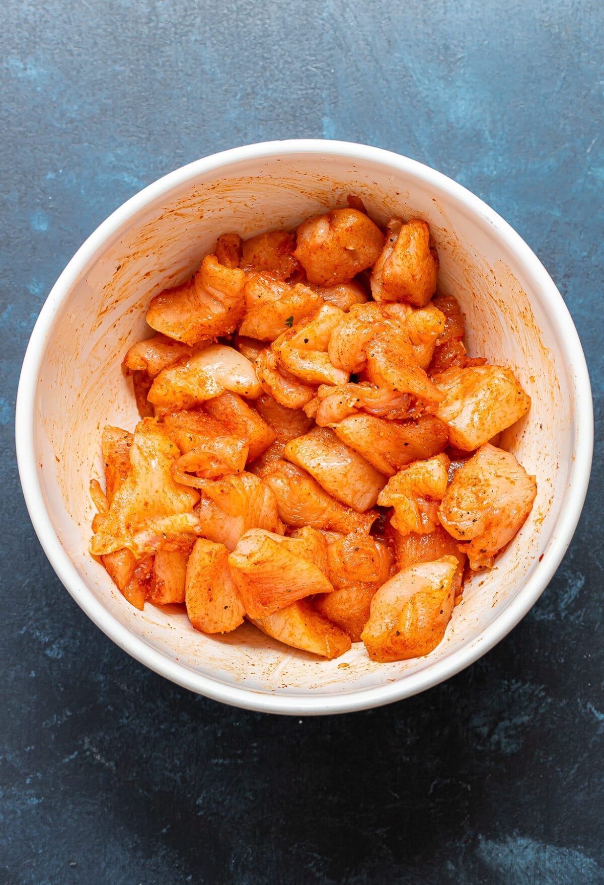 Raw, seasoned chicken pieces in a white bowl on a dark blue textured surface, ready to be cooked for Bang Bang Chicken Bowls. The chicken is coated in orange-colored spices.