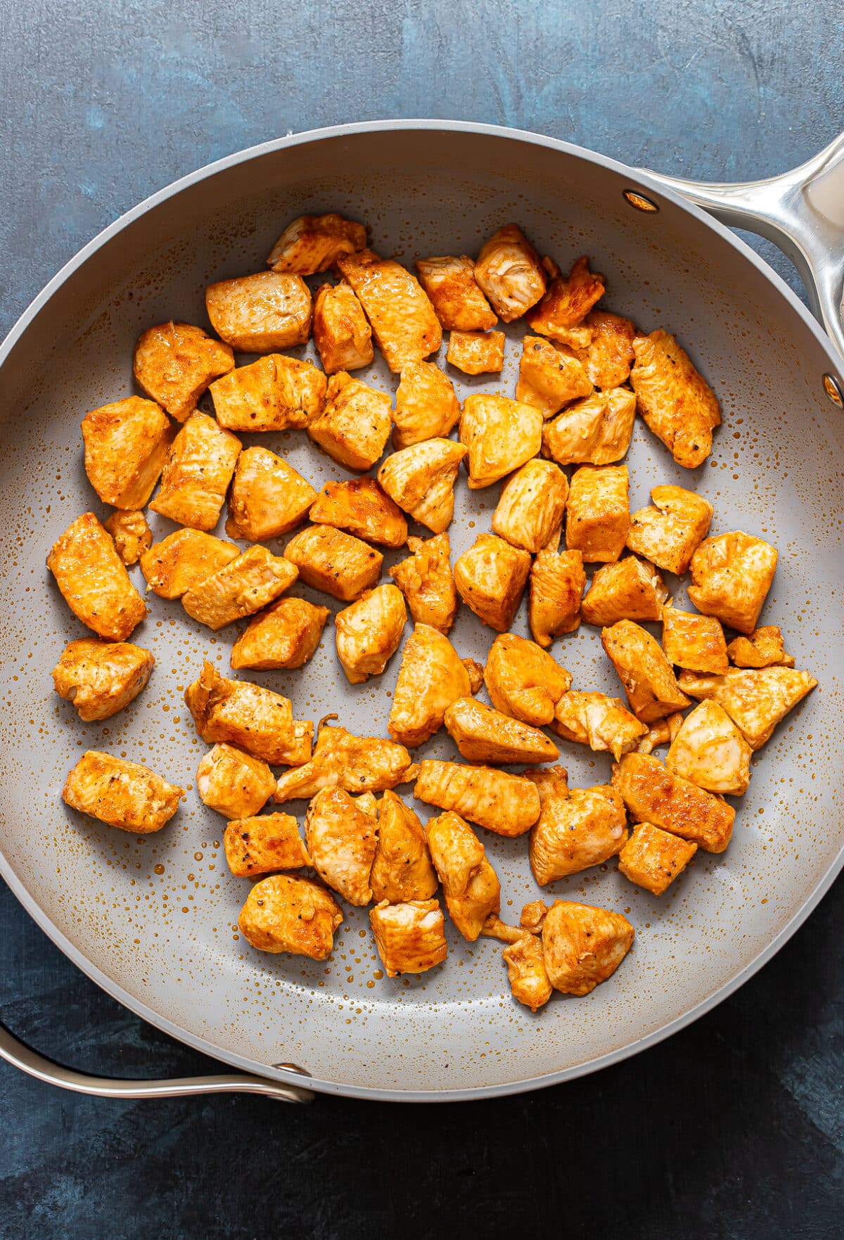 Cooked, seasoned chicken pieces are spread out in a large, round, gray skillet on a dark surface. Perfect for Bang Bang Chicken Bowls, the chicken appears golden brown and lightly crispy, with visible spices on the surface.