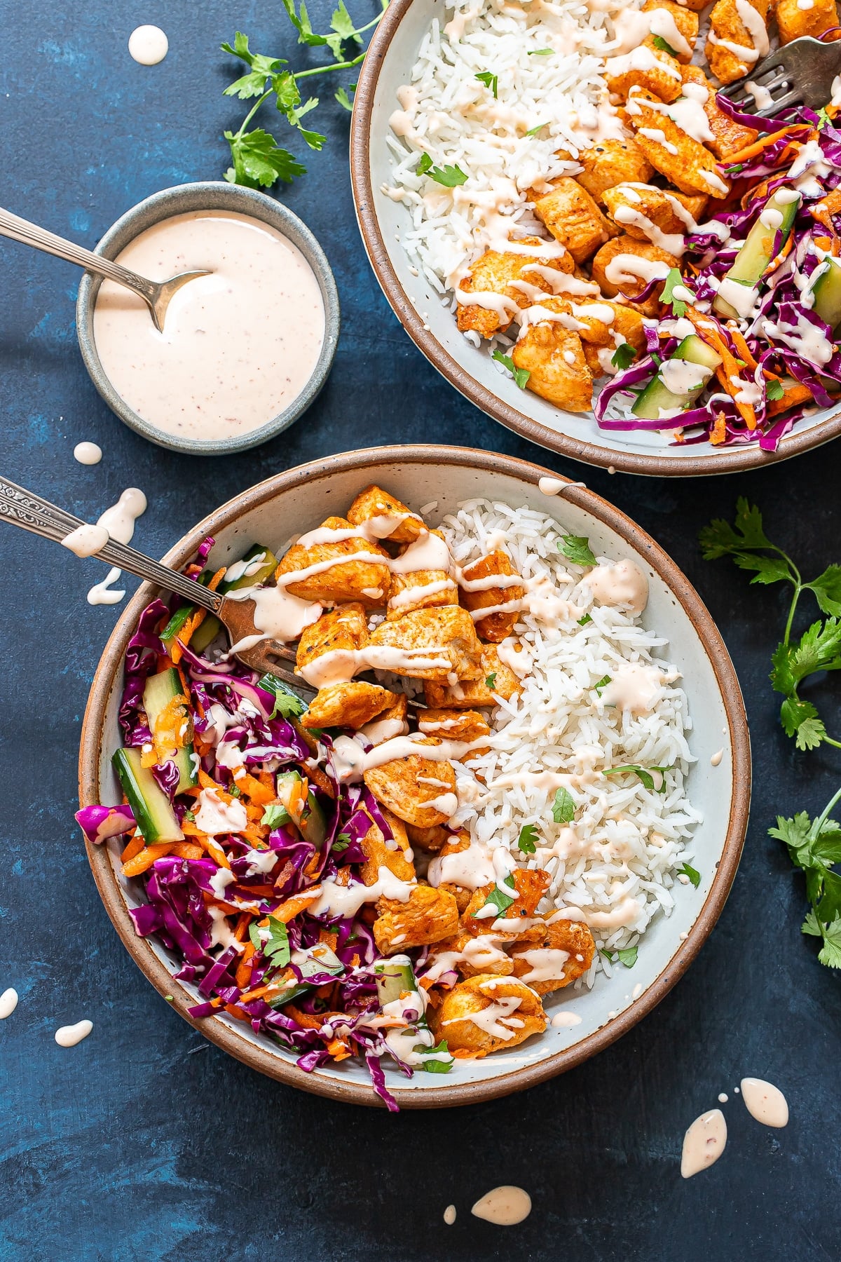 Two Bang Bang Chicken Bowls filled with rice, grilled chicken, purple cabbage slaw, and creamy dressing are placed on a dark blue surface. A small bowl of extra dressing with a spoon sits beside them, while fresh cilantro is scattered around.