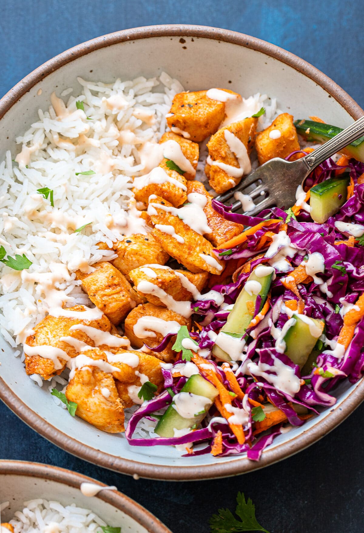 A bowl with white rice, seasoned Bang Bang Chicken pieces, shredded purple cabbage, cucumber, carrots, and a drizzle of creamy dressing. A fork rests in the bowl.