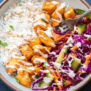 A bowl with white rice, seasoned Bang Bang Chicken pieces, shredded purple cabbage, cucumber, carrots, and a drizzle of creamy dressing. A fork rests in the bowl.