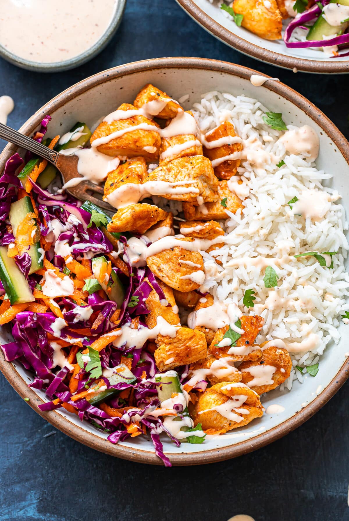 A bowl filled with white rice, seasoned chicken pieces, and a colorful salad of red cabbage, cucumber, and carrots, all drizzled with a creamy sauce—this is the essence of Bang Bang Chicken Bowls. A fork rests in the salad section.