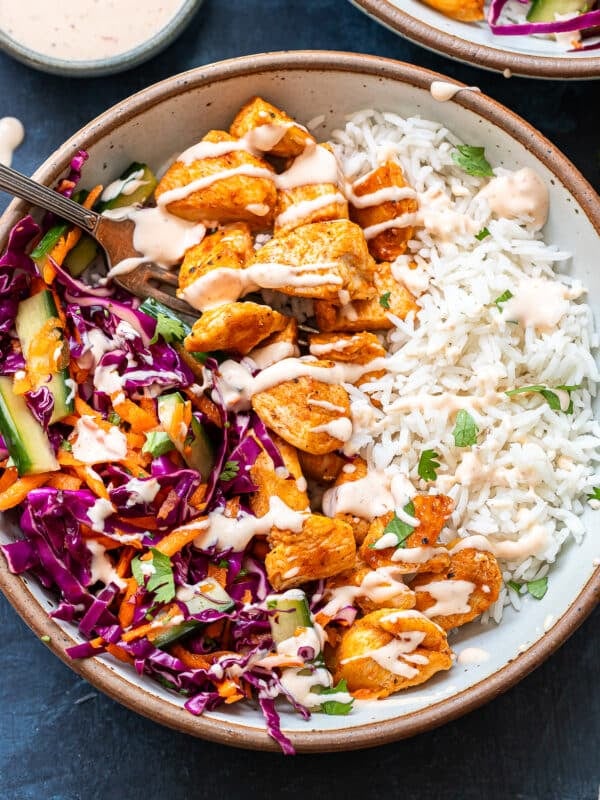 A bowl filled with white rice, seasoned chicken pieces, and a colorful salad of red cabbage, cucumber, and carrots, all drizzled with a creamy sauce—this is the essence of Bang Bang Chicken Bowls. A fork rests in the salad section.