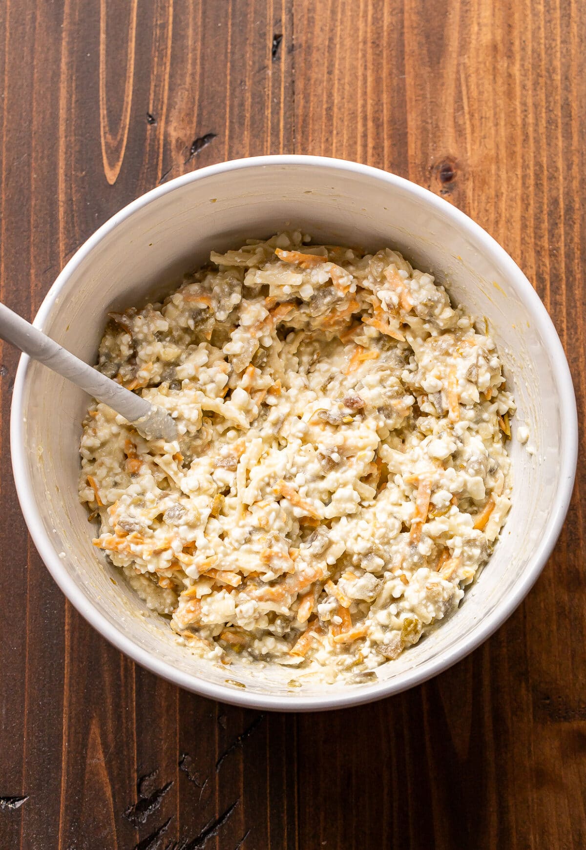 A white bowl with a spoon inside, filled with a mixture of shredded carrots, cottage cheese, and seeds, sits on a wooden surface.