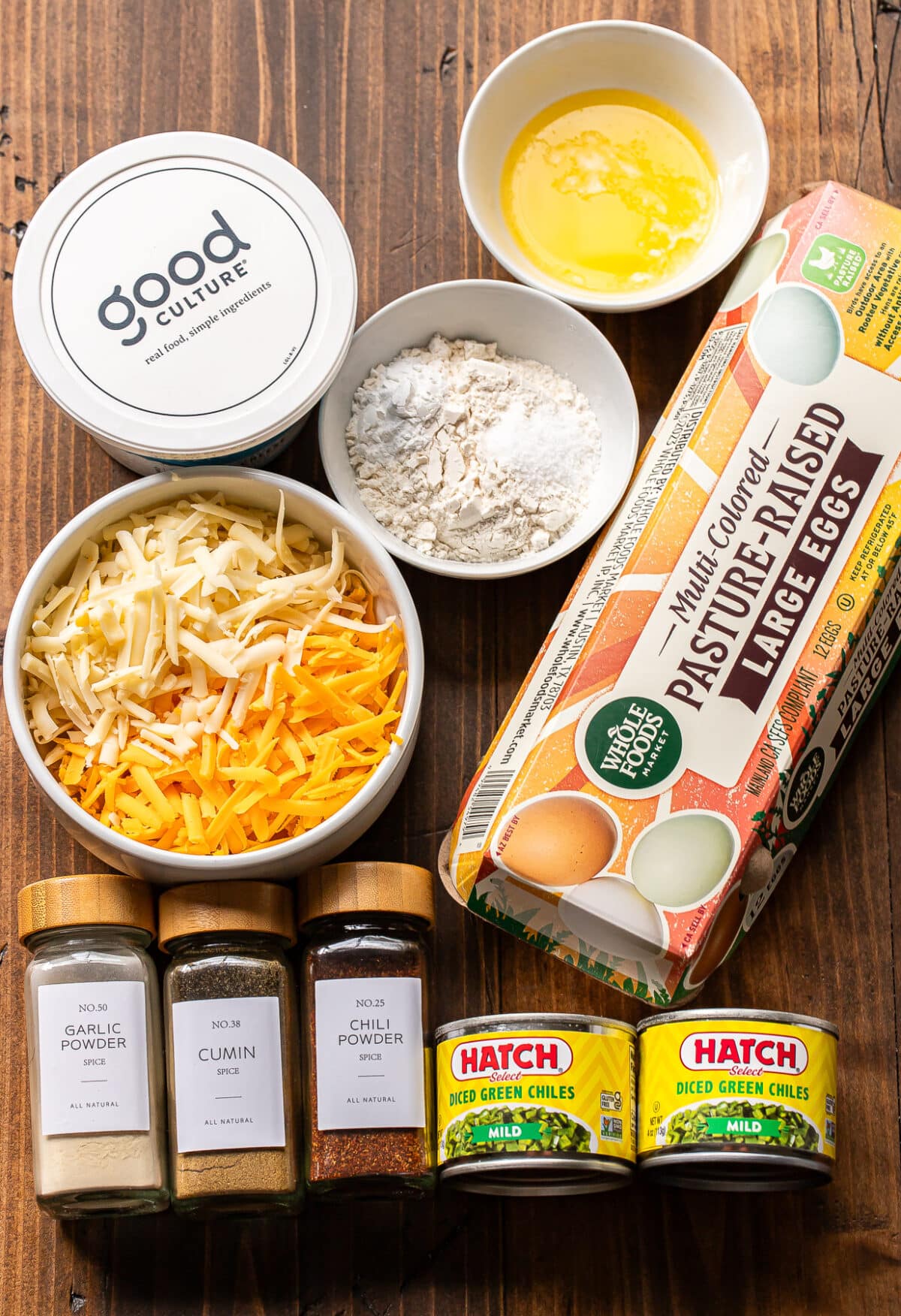 Ingredients for a recipe are laid out, including eggs, shredded cheese, green chilies, cottage cheese, melted butter, flour, plus jars of garlic powder, cumin, and chili powder on a wooden surface.