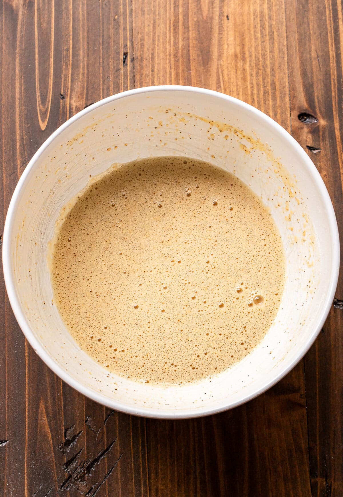White mixing bowl filled with light brown, bubbly batter sits on a wooden surface.