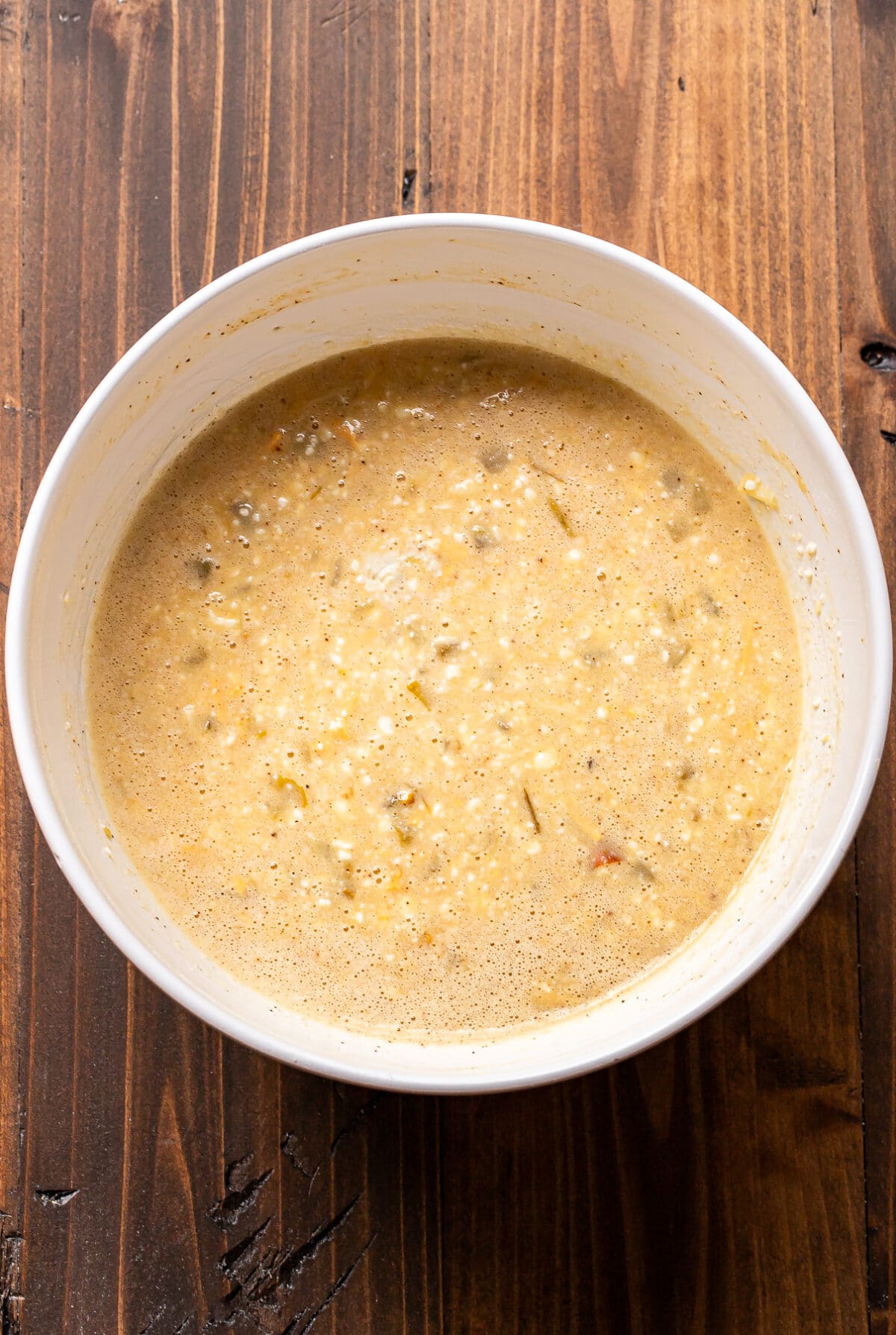 A white bowl filled with a lumpy, beige batter sits on a wooden surface. The mixture appears to contain small bits of vegetables or grains.