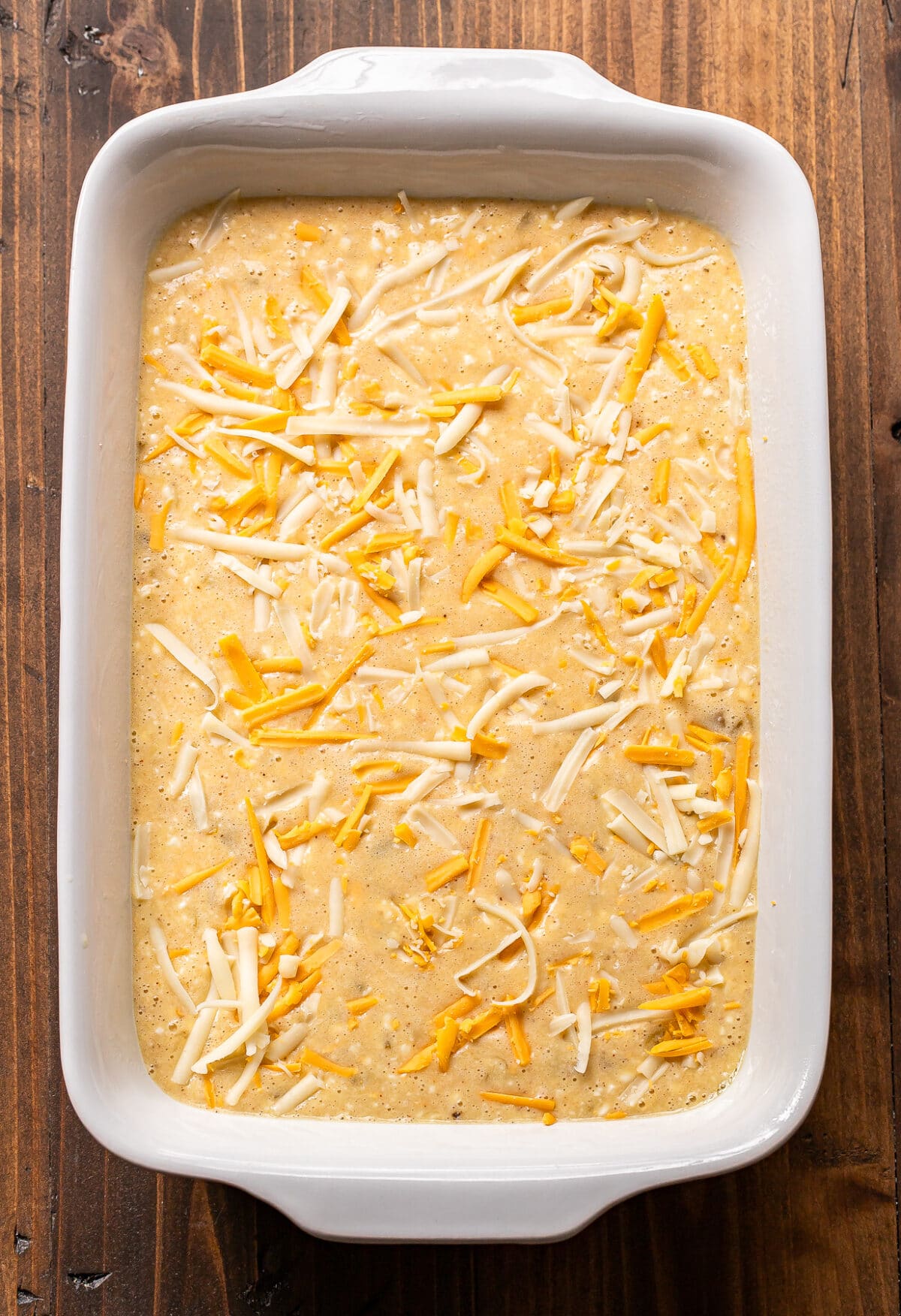 A white baking dish filled with an unbaked casserole mixture, topped with shredded yellow and white cheese, sits on a wooden surface.